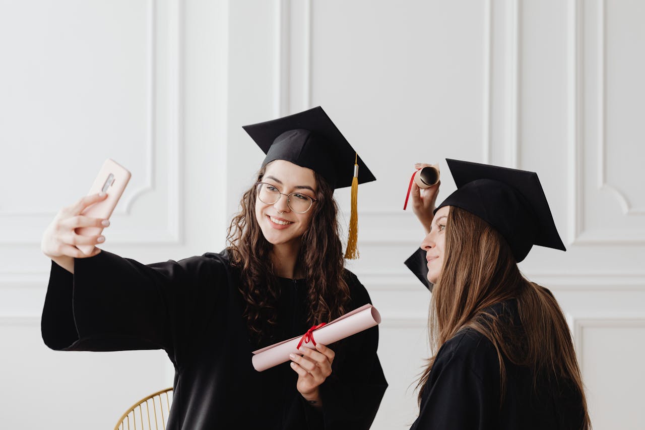 Mastering the First Impression: Your intriguing post title goes here Two happy female graduates in academic gowns taking a selfie while holding diplomas indoors.